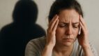Žena drži glavu u bolu, zatvorenih očiju, dok iza nje stoji senka emocionalnog tereta na zidu